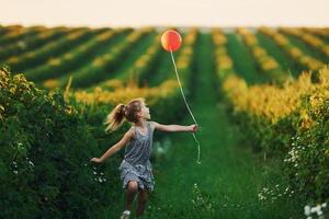 positives kleines mädchen mit rotem ballon in den händen hat spaß auf dem feld zur sommertageszeit foto