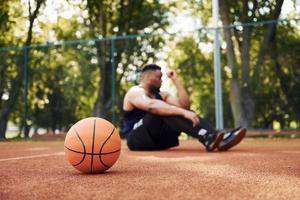 müder afroamerikaner sitzt mit ball auf dem platz im freien auf dem boden. macht eine Pause foto