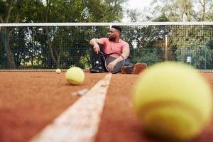 sitzt in der Nähe des Netzes und macht eine Pause. afroamerikanischer mann im rosa hemd sitzt mit tennisschläger auf dem platz im freien foto