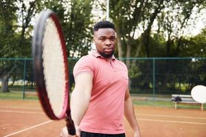 afroamerikanischer Mann in rosafarbenem Hemd posiert für eine Kamera mit Tennisschläger auf dem Platz im Freien foto