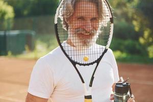 Pause machen und Wasser trinken. Älterer, moderner, stilvoller Mann mit Schläger im Freien auf dem Tennisplatz tagsüber foto