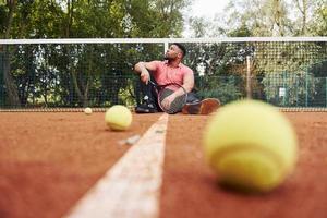 sitzt in der Nähe des Netzes und macht eine Pause. afroamerikanischer mann im rosa hemd sitzt mit tennisschläger auf dem platz im freien foto