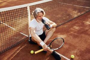 Wasser trinken und auf dem Boden sitzen. Älterer, moderner, stilvoller Mann mit Schläger im Freien auf dem Tennisplatz tagsüber foto