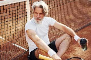 Wasser trinken und auf dem Boden sitzen. Älterer, moderner, stilvoller Mann mit Schläger im Freien auf dem Tennisplatz tagsüber foto