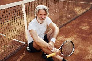 sitzt auf dem Boden und macht Pause. Älterer, moderner, stilvoller Mann mit Schläger im Freien auf dem Tennisplatz tagsüber foto