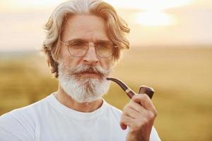 Porträt eines älteren, stilvollen Mannes mit grauem Haar und Bart, der an sonnigen Tagen im Freien auf dem Feld steht und raucht foto
