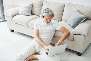 sitzt mit laptop auf dem boden neben dem sofa und hört musik. älterer stilvoller moderner mann mit grauem haar und bart zuhause foto