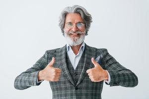 in eleganter Kleidung. stehend gegen weiße Wand. älterer stilvoller moderner mann mit grauem haar und bart zuhause foto