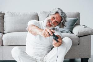 spielt Videospiel mit Controller. älterer stilvoller moderner mann mit grauem haar und bart zuhause foto