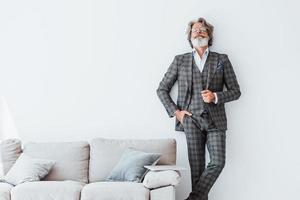 in eleganter Kleidung. stehend gegen weiße Wand. älterer stilvoller moderner mann mit grauem haar und bart zuhause foto