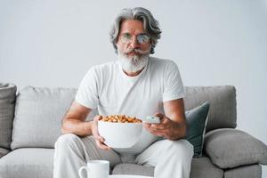 sieht eine Fernsehsendung und isst Popcorn. älterer stilvoller moderner mann mit grauem haar und bart zuhause foto
