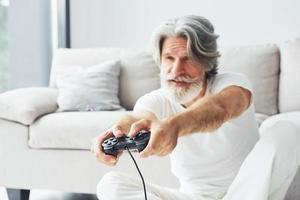 spielt Videospiel mit Controller. älterer stilvoller moderner mann mit grauem haar und bart zuhause foto