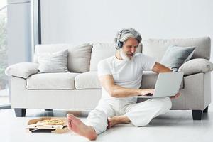 im Kopfhörer. sitzt neben dem Sofa. älterer stilvoller moderner mann mit grauem haar und bart zuhause foto