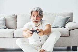 spielt Videospiel mit Controller. älterer stilvoller moderner mann mit grauem haar und bart zuhause foto