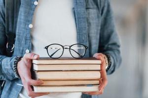Nahaufnahme eines jungen Studenten in Jeanskleidung, der sich im Korridor eines Colleges mit Büchern mit Brillen darauf in den Händen befindet foto