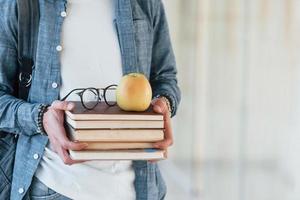 Nahaufnahme eines jungen Studenten in Jeanskleidung, der sich im Korridor eines Colleges mit Büchern mit Apfel darauf in den Händen befindet foto