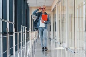 männlicher junger student in jeanskleidung ist im flur eines colleges mit notizblock in den händen