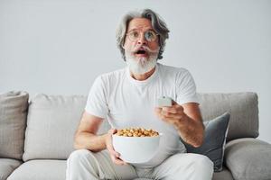 sieht eine Fernsehsendung und isst Popcorn. älterer stilvoller moderner mann mit grauem haar und bart zuhause foto