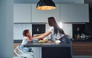 Seitenansicht der Mutter, die mit ihrer kleinen Tochter drinnen in der Küche Essen zubereitet foto