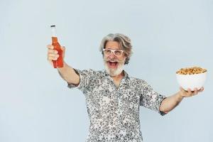 älterer stilvoller moderner mann mit grauem haar und bart zuhause mit flasche getränk und popcorn foto