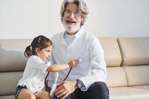 kleines Mädchen, das mit Stethoskop spielt. leitender männlicher arzt mit grauem haar und bart im weißen kittel ist drinnen in der klinik foto