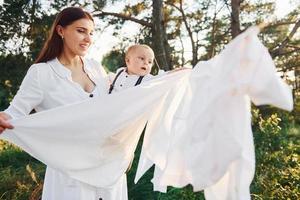 positive hausfrau mit hängt weiße kleidung zum trocknen. junge mutter mit ihrem kleinen sohn ist draußen im wald. schöner Sonnenschein foto