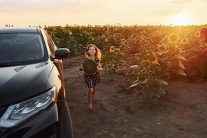 Fröhliches kleines Mädchen geht auf dem landwirtschaftlichen Feld in der Nähe eines modernen schwarzen Autos spazieren und hält Sonnenblumen in der Hand foto