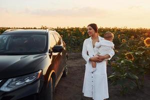 in der nähe eines modernen schwarzen autos. junge mutter mit ihrem kleinen sohn ist draußen im landwirtschaftlichen bereich. schöner Sonnenschein foto