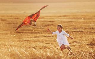 glückliches kleines Mädchen, das im Sommer mit Rotmilan im Freien auf dem Feld läuft foto