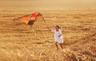 glückliches kleines Mädchen, das im Sommer mit Rotmilan im Freien auf dem Feld läuft foto