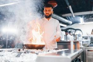 verwendet Bratpfanne. Koch in weißer Uniform, der Essen in der Küche kocht. arbeitsreicher Tag foto