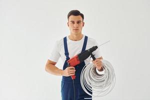 mit bohrer und kabel in den händen. männlicher Arbeiter in blauer Uniform, der im Studio vor weißem Hintergrund steht foto