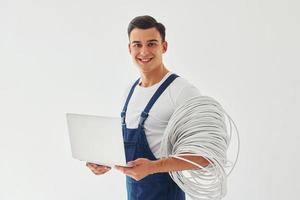 nutzt Laptop. männlicher Arbeiter in blauer Uniform, der im Studio vor weißem Hintergrund steht foto