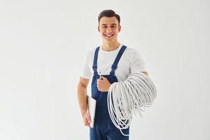 hält Kabel, Notizblock und Schraubendreher. männlicher Arbeiter in blauer Uniform, der im Studio vor weißem Hintergrund steht foto