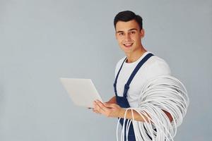 nutzt Laptop. männlicher Arbeiter in blauer Uniform, der im Studio vor weißem Hintergrund steht foto