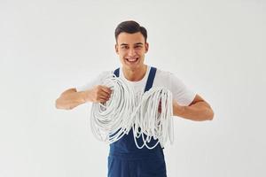 hält Kabel. männlicher Arbeiter in blauer Uniform, der im Studio vor weißem Hintergrund steht foto