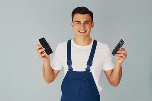 TV-Fernbedienung und Smartphone. männlicher Arbeiter in blauer Uniform, der im Studio vor weißem Hintergrund steht foto