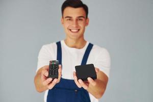 TV-Fernbedienung und Smartphone. männlicher Arbeiter in blauer Uniform, der im Studio vor weißem Hintergrund steht foto