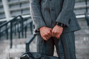 hält Tasche in den Händen. Junger Geschäftsmann in grauer Abendgarderobe ist draußen in der Stadt foto