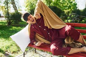 im Park auf der Bank liegen. junger mann im pyjama ist draußen auf der straße. fühlt sich schläfrig foto