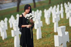 mit Blumen in den Händen. junge Frau in schwarzer Kleidung, die den Friedhof mit vielen weißen Kreuzen besucht. Vorstellung von Begräbnis und Tod foto