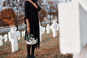 mit Blumen in den Händen. junge Frau in schwarzer Kleidung, die den Friedhof mit vielen weißen Kreuzen besucht. Vorstellung von Begräbnis und Tod foto