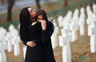 sich umarmen und weinen. Zwei junge Frauen in schwarzer Kleidung besuchen den Friedhof mit vielen weißen Kreuzen. Vorstellung von Begräbnis und Tod foto