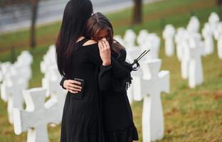 sich umarmen und weinen. Zwei junge Frauen in schwarzer Kleidung besuchen den Friedhof mit vielen weißen Kreuzen. Vorstellung von Begräbnis und Tod foto