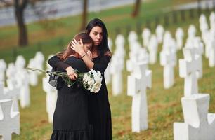 sich umarmen und weinen. Zwei junge Frauen in schwarzer Kleidung besuchen den Friedhof mit vielen weißen Kreuzen. Vorstellung von Begräbnis und Tod foto