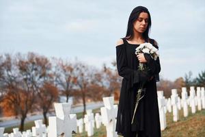 mit Blumen in den Händen. junge Frau in schwarzer Kleidung, die den Friedhof mit vielen weißen Kreuzen besucht. Vorstellung von Begräbnis und Tod foto