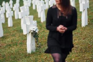 gibt Respekt, indem er Blumen legt. junge Frau in schwarzer Kleidung, die den Friedhof mit vielen weißen Kreuzen besucht. Vorstellung von Begräbnis und Tod foto