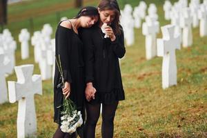 hält Blumen. Zwei junge Frauen in schwarzer Kleidung besuchen den Friedhof mit vielen weißen Kreuzen. Vorstellung von Begräbnis und Tod foto