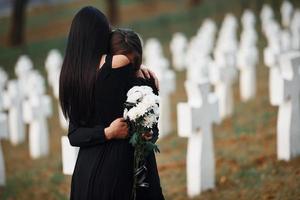 sich umarmen und weinen. Zwei junge Frauen in schwarzer Kleidung besuchen den Friedhof mit vielen weißen Kreuzen. Vorstellung von Begräbnis und Tod foto