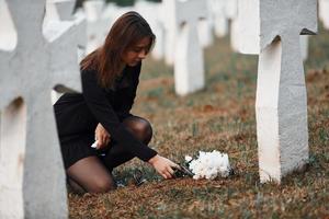 gibt Respekt, indem er Blumen legt. junge Frau in schwarzer Kleidung, die den Friedhof mit vielen weißen Kreuzen besucht. Vorstellung von Begräbnis und Tod foto